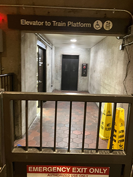 A picture of the pathway to the elevator that goes to the Upper Platform of the station. In the foreground there is the emergency exit gate for the pathway. Further back, there are two yellow wet floor cones on the right, an open storage room on the left, and the elevator straight back.